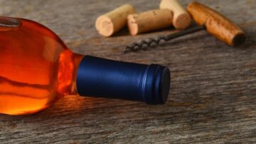 زجاجة نبيذ وردي مع غطاء أزرق، موضوعة على سطح خشبي، مع عدة سدادات وفتاحة زجاجات في الخلفية.