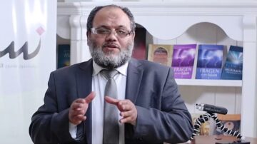 رجل يرتدي بدلة رمادية وقميص أبيض، يتحدث أمام مكتبة تحتوي على كتب دينية. يظهر خلفه شعار مشروع إسلامي. يعبّر عن أفكاره بلغة واضحة وإيماءات يدوية.