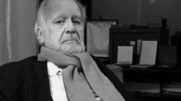 رجل مسن يجلس على أريكة مع تعبير جاد، يرتدي سترة داكنة وشال رمادي. تظهر خلفه أشياء مكتبية غير واضحة في بيئة داخلية.