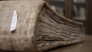 كتاب قديم مع غلاف جلدي ومظهر متهالك، يظهر حوافه البالية وصفحاته المتجعدة. الخلفية تحتوي على رفوف مكتبة تحتوي على كتب أخرى.