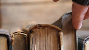 يد تظهر وهي تلمس حواف كتب قديمة موضوعة بجانب بعضها، مع تركيز على تفاصيل الغلاف والتآكل. خلفية خشبية تضيف جوًا دافئًا للترتيب.