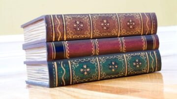 كتب قديمة ملونة موضوعة بشكل عمودي على أرضية خشبية، تتكون من ثلاثة مجلدات بتصاميم مزخرفة.