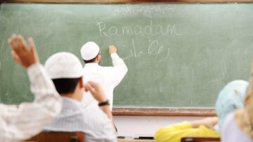 معلم يكتب كلمة رمضان على السبورة الخضراء في فصل دراسي، بينما يرفع الطلاب أيديهم للمشاركة. تعكس الصورة أجواء التعليم والتفاعل خلال شهر رمضان.