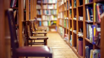 مكتبة تحتوي على رفوف مليئة بالكتب، مع صف من الكراسي الخشبية على جانب الممر. البيئة هادئة ومناسبة للدراسة والقراءة.