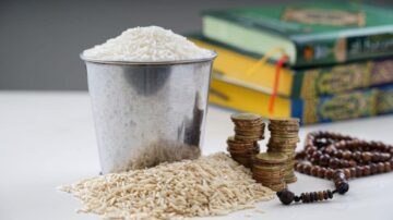 أرز أبيض في دلو معدني مع حبوب أرز على السطح. بجانبها عملات معدنية مكدسة وسجادة صلاة، مع كتب دينية في الخلفية. تعكس الصورة موضوعات الزراعة والاقتصاد والتقاليد الدينية.