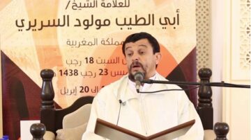 رجل يتحدث في جلسة علمية، يرتدي الزي التقليدي المغربي، مع كتاب مفتوح في يده. خلفية الجلسة تتضمن نصوص باللغة العربية تشير إلى موضوع المحاضرة وتاريخها.