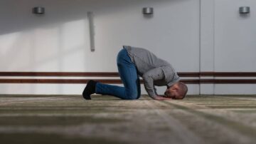 رجل يرتدي قميصًا مخططًا وزوجًا من الجينز، يركع على الأرض في وضعية سجود، مع التركيز على تعبيراته الجادة، في بيئة داخلية ذات إضاءة طبيعية.