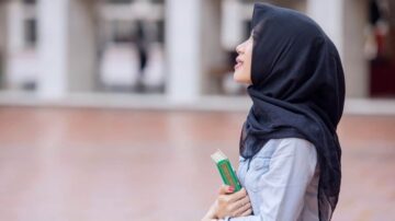 امرأة ترتدي حجابًا أسود، تحمل كتابًا بين يديها، وتنظر إلى السماء بتفكير. تظهر في خلفية مشوشة لمكان مفتوح.