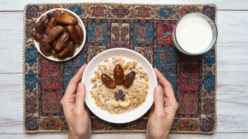 طبق من الحبوب مع التمر والمكسرات، يُحمل بيدين، بجانب كوب من الحليب وصحن من التمر، على سجادة مزخرفة. وجبة صحية وغنية بالعناصر الغذائية.