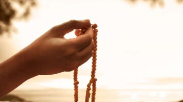 يد تمسك بسبحة مصنوعة من حبات خشبية، مع خلفية ضبابية تضفي جوًا من الهدوء والسكينة. تبرز السبحة كرمز للتأمل والروحانية.