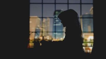 امرأة محجبة تجلس بجوار نافذة كبيرة، silhouetted against منظر حضري ليلي. يظهر في الخلفية أضواء المدينة وناطحات السحاب، مما يخلق جوًا هادئًا وتأمليًا.