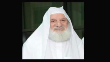 رجل مسن ذو لحية بيضاء، يرتدي غطاء رأس أبيض وثوب تقليدي. يبدو مبتسمًا ويقف أمام خلفية داكنة.