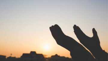 أيد مرفوعة نحو السماء خلال غروب الشمس، مع وجود ظلال واضحة للمنازل في الخلفية. تعبر الصورة عن الأمل والتأمل والسكينة.