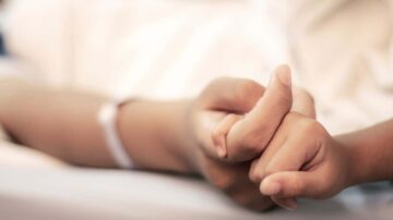 يدين متشابكتين، إحداهما تحمل سوارًا طبيًا، في بيئة مستشفى. تعكس الصورة مشاعر الدعم والراحة.