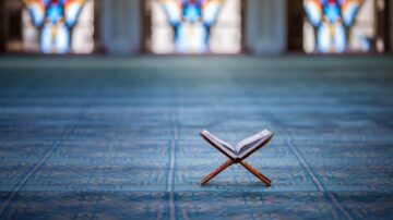 كتاب مفتوح على حامل في مركز مسجد، مع أرضية زرقاء مزخرفة ونوافذ ملونة في الخلفية.