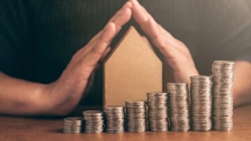 رجل يحمي نموذجاً لمنزل بيديه، بينما تتكدس عملات معدنية مرتبة في صفوف متزايدة، مما يشير إلى ادخار المال والاستثمار في العقارات.