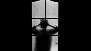 شخص يقف خلف باب زجاجي مغطى بالندي، يعبر عن الإحباط أو القلق من خلال وضع يديه على رأسه، مما يخلق صورة دراماتيكية تعكس المشاعر العميقة.