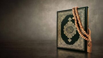 قرآن كريم مفتوح مع مسبحة خشبية، يظهر بتفاصيل دقيقة على خلفية رمادية داكنة.