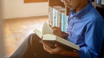 رجل يرتدي قميصًا أزرق يجلس على الأرض ويقرأ كتابًا في مكتبة. في الخلفية، توجد رفوف مليئة بالكتب، والإضاءة الطبيعية تضفي جوًا هادئًا.