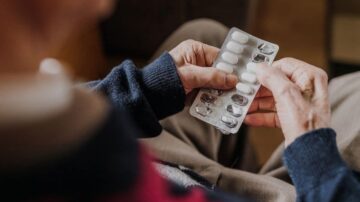 رجل مسن يجلس على أريكة، ممسكًا بشريط من الحبوب، يظهر التركيز على وجهه. الخلفية غير واضحة، مما يبرز اهتمامه بالأدوية.