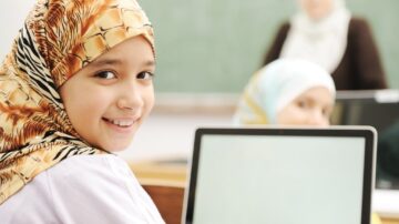 فتاة ترتدي حجاباً ملوناً تجلس أمام كمبيوتر محمول مبتسمة في فصل دراسي. في الخلفية، معلمة ترتدي حجاباً وتظهر بعض الطالبات الأخريات. الصورة تعكس بيئة تعليمية شاملة.