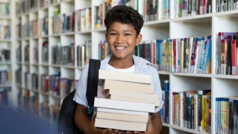 فتى صغير يحمل مجموعة كتب وتظهر خلفة مكتبة كبيرة مليئة بالكتب