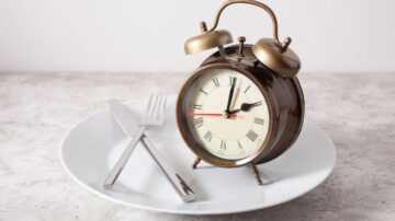 ساعة منبه معدنية قديمة على طبق أبيض مع سكين وشوكة. الساعة تشير إلى وقت الوجبة، مما يرمز إلى أهمية توقيت الطعام. تصميم أنيق يجمع بين الأناقة والكلاسيكية.