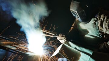 لحام شخص يرتدي خوذة واقية ومئزر، يعمل على قطعة معدنية، مع انبعاث شرارات وضوء ساطع. يُظهر المشهد عملية اللحام في ورشة عمل، مما يبرز أهمية السلامة في الحرف اليدوية.