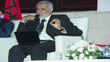 رجل يتحدث في مؤتمر، يحمل ميكروفونًا ويجلس على كرسي أبيض. خلفه علم المغرب، أمامه لاب توب وزهور بيضاء.