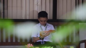 ولد يجلس على مقعد في منطقة مظللة، يقرأ كتابًا. تظهر خلفه نباتات خضراء، مع نافذة ذات ستائر. يُظهر التركيز والانغماس في القراءة.