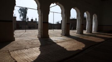 منظر داخلي يظهر أقواسًا معمارية بيضاء تخلق ظلالًا على الأرضية. السطح مغطى بسجاد، والخلفية تحتوي على معالم معمارية تقليدية. يضيء الضوء الطبيعي المكان، مما يبرز التفاصيل المعمارية.