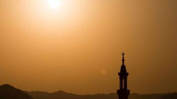 صورة ظلية لمئذنة مسجد مع غروب الشمس في الخلفية، تظهر الأفق الجبلي بلون برتقالي دافئ.