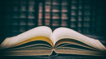 كتاب مفتوح على طاولة، يظهر الصفحات الداخلية بلون كريمي. الخلفية ضبابية تحتوي على رفوف كتب مظلمة، مما يضيف جوًا هادئًا ومناسبًا للقراءة.