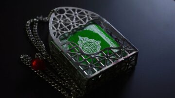 تصميم فضي مزخرف يحمل كتابًا أخضر، يمثل القرآن. يتضمن سلسلة فضية مع خرزة حمراء، ويظهر على سطح أسود.