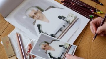 صورة تُظهر شخصًا يرسم بورتريه باستخدام أقلام ملونة، مع وجود صورة مرجعية ملصقة بجانب الرسمة. الخلفية خشبية، وتظهر أدوات الرسم مثل أقلام التلوين وبعض المواد الفنية.