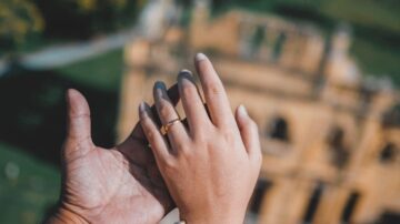 يد تحمل خاتم زفاف، مع يد أخرى تحتها، وخلفية ضبابية لمبنى تاريخي.