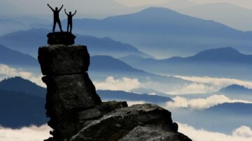 Two silhouetted figures stand triumphantly on a rocky summit, surrounded by layers of misty blue mountains. The scene captures a sense of adventure and accomplishment against a serene backdrop of clouds and peaks.