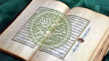 صفحة مفتوحة من كتاب قديم مكتوب بخط عربي جميل، تحتوي على نصوص دينية وزخارف تقليدية، مع خلفية خضراء.