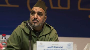 رجل يرتدي قبعة تقليدية ولها لون زيتوني، يجلس خلف ميكروفون مع تعبير جدي. خلفه لافتة تحتوي على نص عربي. يُظهر المشهد مناسبة رسمية أو مؤتمر.