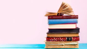 مجموعة من الكتب المكدسة، تشمل كتاب مفتوح في الأعلى وكتب ملونة بأحجام مختلفة بالأسفل، مع خلفية وردية. تعكس الصورة حب القراءة والثقافة.