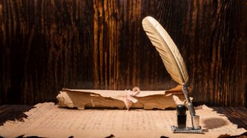 قلم ريشي ذهبي بجانب حبر أسود، موضوعة على ورقة قديمة. خلفية خشبية داكنة تضيف طابعًا تاريخيًا.