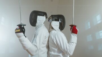 صورة لرياضيين متنافسين في رياضة المبارزة يقفان背向背، مرتديين خوذات وأزياء المبارزة البيضاء. يحمل كل منهما سلاح المبارزة، مما يعكس التحدي والتركيز في هذه الرياضة. الخلفية ضبابية، مما يضيف جوًا دراماتيكيًا للمشهد.