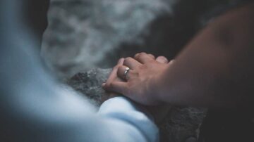 يدين متشابكتين مع خاتم زفاف، تظهر اللحظة الحميمة بين شخصين. الخلفية غير واضحة، مما يبرز التركيز على الأيدي.