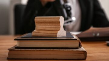 مطرقة خشبية على مجموعة من الكتب القديمة، مع شخص يرتدي بدلة خلفها يحمل ميكروفون. تعكس الصورة أجواء قانونية أو مهنية.