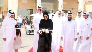 افتتاح حدث بمشاركة مجموعة من الأشخاص يرتدون الملابس التقليدية، مع امرأة في المقدمة تقطع شريط الافتتاح. يظهر في الخلفية مجموعة من الرجال والنساء، يتواجدون في موقع خارجي مزين بالسجاد الأحمر.