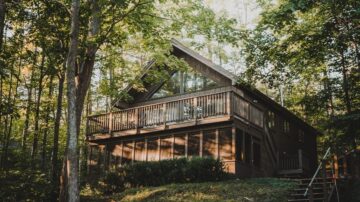 منزل خشبي حديث محاط بالأشجار، يظهر بوضوح تصميمه الزجاجي الكبير. يحتوي على شرفة أمامية ومساحة خضراء، مما يبرز جمال الطبيعة المحيطة.