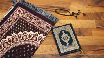 سجادة صلاة مزخرفة بلون بني مع مصحف مفتوح بجانبها، وسبحة خشبية. الخشب في الخلفية يضيف لمسة دافئة.