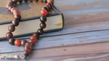 سبحة ملونة مصنوعة من الخرز، موضوعة على كتاب ذو غلاف مزخرف. يظهر الكتاب بلون أزرق مع خشب طبيعي، بينما تعكس الخرزات ألوانًا دافئة مثل الأحمر والبني.