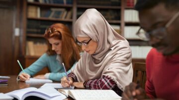 طالبات يدرسن في مكتبة، واحدة ترتدي حجابًا وتكتب في دفتر، بينما الأخريين يكتبن أيضًا. الخلفية تحتوي على رفوف مليئة بالكتب.