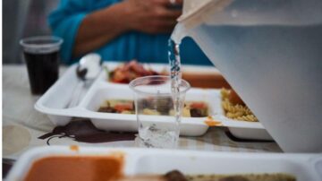 صورة تُظهر شخصًا يصب الماء من وعاء إلى كوب شفاف، مع أطباق طعام متنوعة على الطاولة. يشمل المشهد أطباقًا تحتوي على المعكرونة والصلصة، إلى جانب مشروب غازي في كوب آخر.